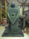 3m bronze sculpture. Stylised adult standing; arms round child, both facing us. Green-grey patina. On plinth. In the foundry.