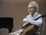 Still image from TV Masterclass. To the left is the back of a black grand piano. Tortelier is facing left with his cello in the crook of his left arm, talking animatedly.