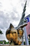 Bronze dagger sculpture with gold handle from the ground. Arches behind, with large Malaysian flag above. Sunny with clouds.