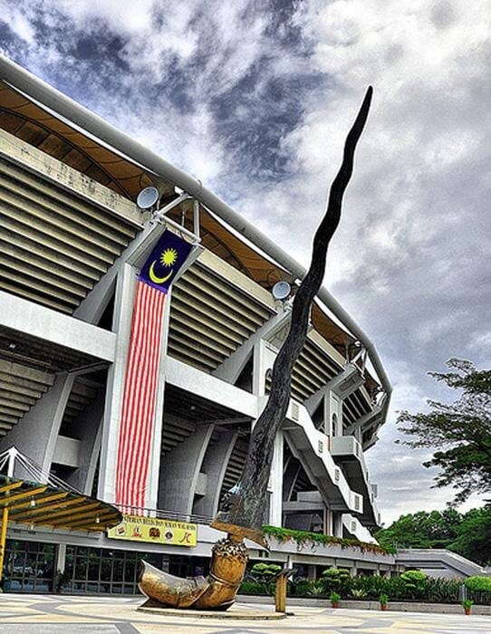 Bronze dagger sculpture with gold handle. Stadium behind, draped in a huge Malaysian flag pennant. Sunny.