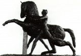 Photo of bronze prancing horse sculpture being restrained by two naked men, one each side, gripping the halter. In black modelling clay with a timber support at the front. Left side.