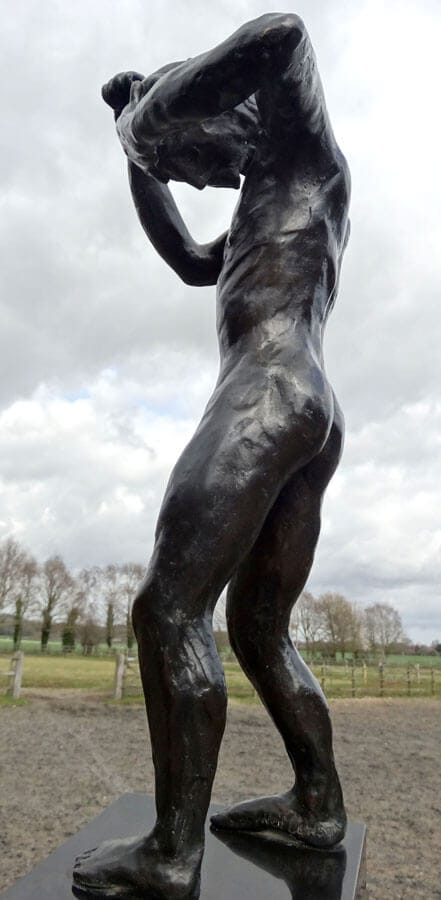 This colour photo shows a fine art bronze sculpture called Speak to us of Crime and Punishment. It is a an adult human with a male physique standing. He is cowering in shame. His head is bent forward so that his chin is almost touching his chest. His left arm is raised horizontally with his left hand covering his forehead. Both legs are slightly bent at the knee. The right hand and arm areup and bent in a defensive, punching posture. He is standing on a highly-polished stone plinth in a brown field. In the middle distance is a wooden post and rail fence with an open gateway to the left of the sculpture. The field beyond has aging grass in it and its boundary is another wooden post and rail fence with immediately behind a row of leafless tall trees, several with ivy climbing the trunks. There are hills in the far distance and the sky is almost completely full of fluffy pal grey and white clouds. We are looking at the sculpture's left side and he is therefore facing left.