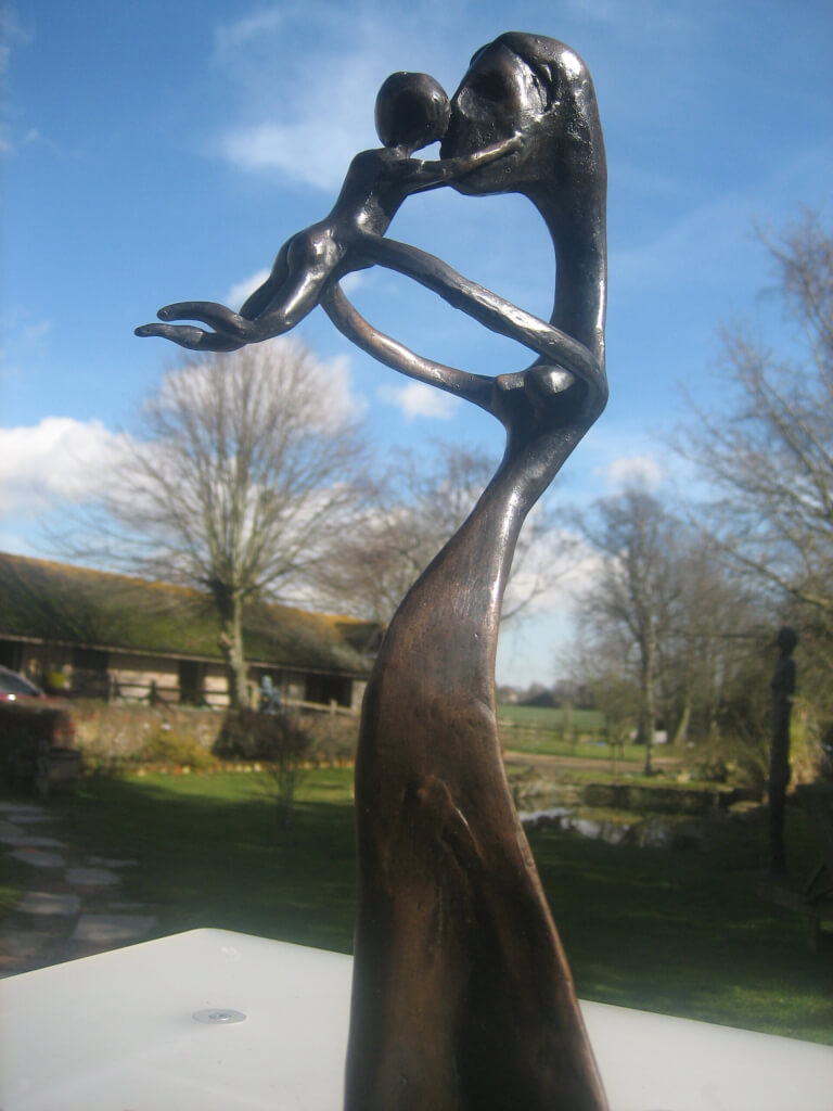 A Joyous Moment - Fine Art Bronze of a mother standing, arms outstretched, holding a 'flying' baby and kissing its head.