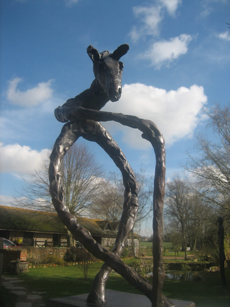 A Man Called Horse - Sculpture. This colour photo shows a fine art bronze sculpture outdoors. The sculpture is of a semi-abstract standing figure and the photo is taken from the front. It is standing on two legs - possibly back legs and the right one is crossed across the front of the left. It has a very short body and from the right shoulder is an elongated limb sloping forwards and down at about 30 degrees to the horizontal. This limb (it is difficult to decide if it is an arm or a foreleg of a horse) makes a sudden turn and drops vertically to the ground below the two feet of the back legs. It could be a hand holding a stick or an anatomically unusual joint. There is no left arm or foreleg. Beyond the shoulder is a horse's neck which continues upwards at 30 degrees and then bends downwards, at 45 degrees culminating in a horse's head with two ears pricked forward. The bronze shows the daubs of clay and is a traditional natural bronze colour. The background shows a garden, with a green lawn a pond a crazy-paving path and a 1m high brick boundary wall. Behind the wall is a long, low, single storey building and there are some mature trees. The sun is out and there is a blue sky with several fluffy white clouds. Beyond the single-storey building are gree fields stretching away into the distabce.