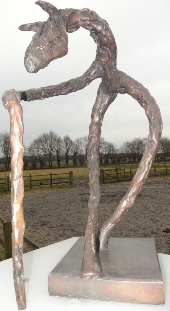 A Man Called Horse - Sculpture. This colour photo shows a fine art bronze sculpture outdoors. The sculpture is of a semi-abstract standing figure and the photo is taken from the front. It is standing on two legs - possibly back legs.. It has a very short body and from the right shoulder is an elongated limb sloping forwards and down at about 30 degrees to the horizontal. This limb (it is difficult to decide if it is an arm or a foreleg of a horse) makes a sudden turn and drops vertically to the ground below the two feet of the back legs. It could be a hand holding a stick or an anatomically unusual joint. There is no left arm or foreleg. Beyond the shoulder is a horse's neck which continues upwards at 30 degrees and then bends downwards, at 45 degrees culminating in a horse's head with two ears pricked forward. The bronze shows the daubs of clay and is a traditional natural bronze colour. The background shows a brown field with post and rail fencing and beyond that green fields, also with post and rail fencing. One of the fences has a row of tall trees. Behind the trees are some low, The sky is grey and quite dull.