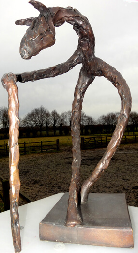 A Man Called Horse - Sculpture. This colour photo shows a fine art bronze sculpture outdoors. The sculpture is of a semi-abstract standing figure and the photo is taken from the front. It is standing on two legs - possibly back legs.. It has a very short body and from the right shoulder is an elongated limb sloping forwards and down at about 30 degrees to the horizontal. This limb (it is difficult to decide if it is an arm or a foreleg of a horse) makes a sudden turn and drops vertically to the ground below the two feet of the back legs. It could be a hand holding a stick or an anatomically unusual joint. There is no left arm or foreleg. Beyond the shoulder is a horse's neck which continues upwards at 30 degrees and then bends downwards, at 45 degrees culminating in a horse's head with two ears pricked forward. The bronze shows the daubs of clay and is a traditional natural bronze colour. The background shows a brown field with post and rail fencing and beyond that green fields, also with post and rail fencing. One of the fences has a row of tall trees. Behind the trees are some low, The sky is grey and quite dull.