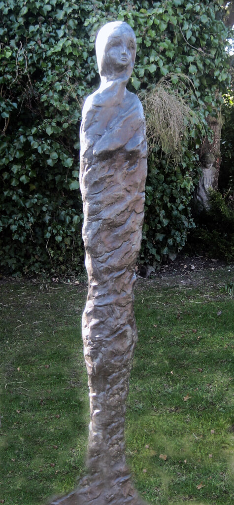 A tall, thin bronze sculpture of a standing woman wrapped in a shroud from head to toe, with only her delicate face showing.