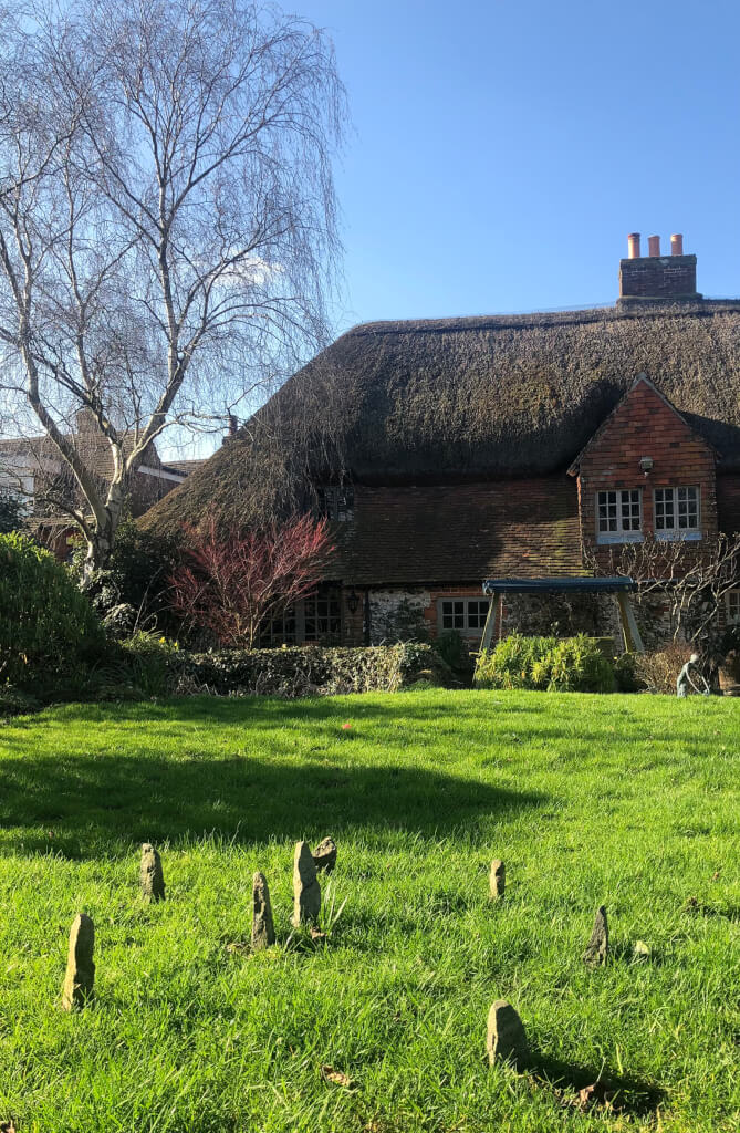 This is a colour photo of a lawn, not recently mown, with a small stone circle in the foreground. The top half of the photo shows a thatched roof above a brick-and-flint cottage with a tile-hung gable end housing white-painted casement windows. To the left of the cottage is a tall silver birch tree which is not yet in leaf. In front of the cottage is a low stone wall and some low shrubs. At the back of the lawn is a small bronze sculpture of a standing human, stooping forwards with arms outstretched. There are 6 stones forming the circle. They are tall (the tallest is ~400mm) and thin and yellowish - possibly sandstone. The centre of the circle is marked by two slightly taller stones. The sun is out and shining from the left. There is a large tree shadow, immediately behind the circle on the lawn. The sky above the thatched roof and to the left of the cottage is a bright, pale blue and almost completely cloudless.