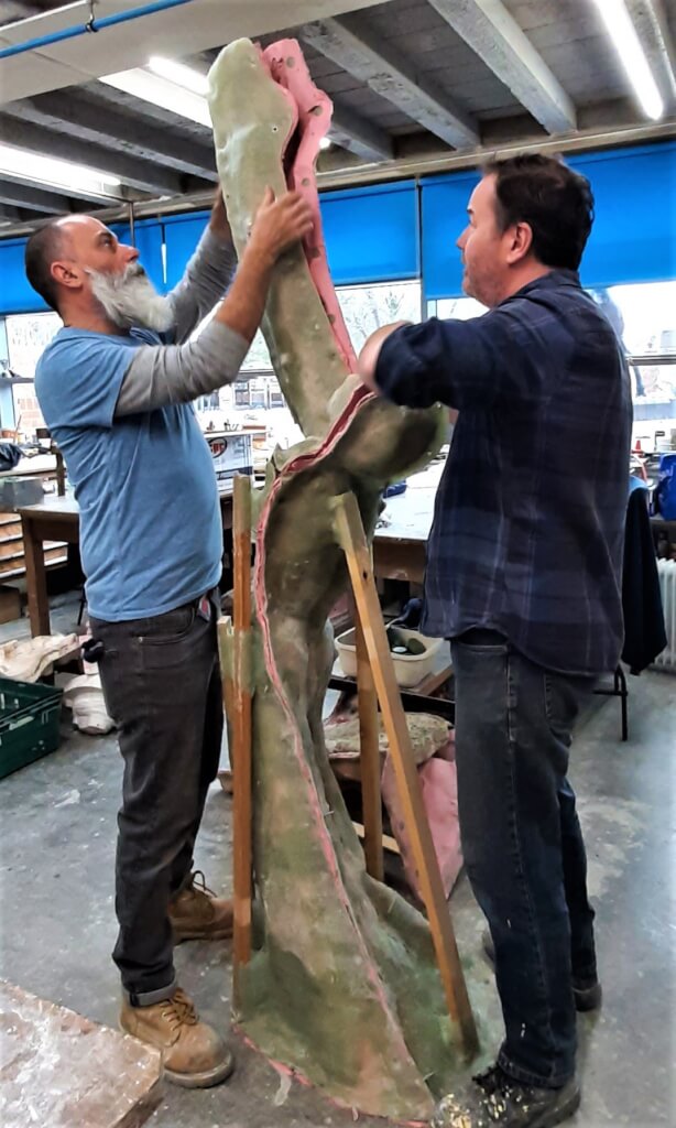 Two men facing each other in a workshop. Between them is a mould made from the clay model enlarged from the original. The mould is made in sections and the men are dismantling it ready for the next part of the process.