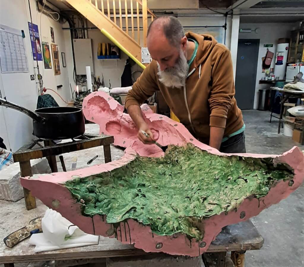 Speak to us of Freedom sculpture. A man in a workshop. He's bending over a large pink object which is a section of silicone mould of the clay model and he is 'painting' green wax into the mould from a large saucepan