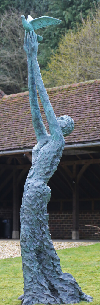 A exaggeratedly tall standing female figure with long skirts. Her arms are held aloft and she is releasing a dove. The bronze statue is standing on a green grass lawn in front of an open barn-type building.