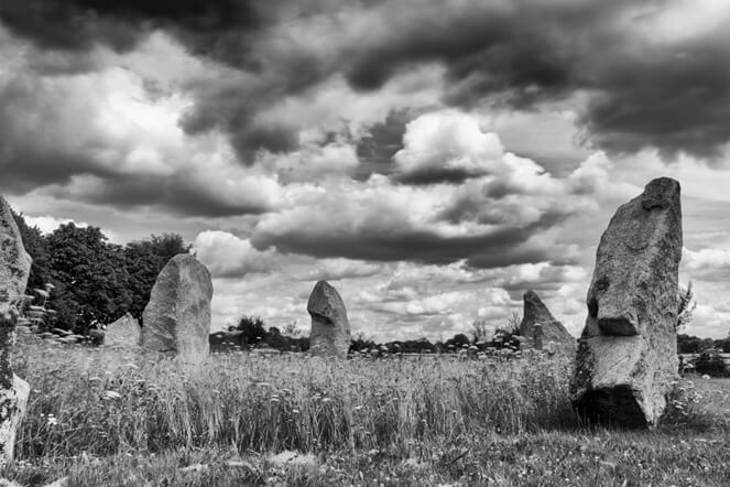 This black & white photo is taken at ground level. It shows six standing stones in a field. The stones seem to form a cluster. The grass in the field has been cut short outside the cluster, but left uncut in the centre of the cluster. There is a very heavy, atmospheric sky, full of clouds of strongly contrasting whites and greys. The tree-lined horizon is just visible behind most of the stones, but the left most ones have oak trees in full leaf close by behind them.