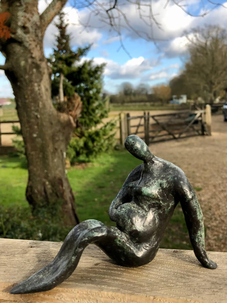 A semi-abstract cast bronze figure, sitting on the ground, legs together, feet on the ground and knees raised. Her left arm is out behind her to keep her upright and she is very heavily pregnant. Her right hand rests on her tummy as if feeling for the baby kicking.