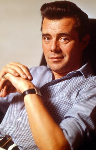 This is a colour photo of Dirk Bogarde sitting in a chair. He is leaning backwards quite far, with both elbows resting on the arms of the chair and his hands meeting in a form of arch in front of him. His right hand is over the top of his left, which has a gold signet ring on his little finger. He is wearing a square wristwatch with a black leather strap. He is lightly tanned. He is looking straight at the lens and has a half-smile. He has very dark hair and eyebrows and dark brown eyes.