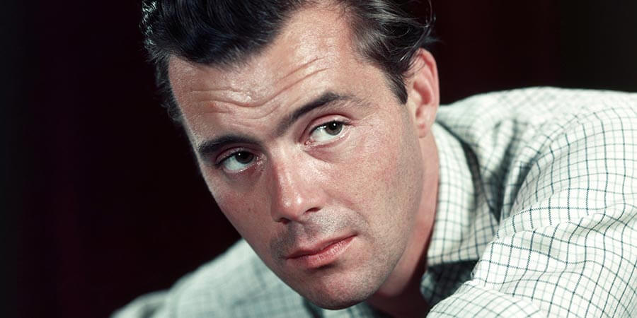 This is a colour photograph of Dirk Bogarde. It shows his face and left shoulder which are in sharp focus, but his neck and right shoulder are out of focus. His body is leaning forward from right to left and his head is turned to the left towards the camera and slightly beyond. He is wearing a white shirt with a dark blue and pale grey check formed by lines crossing at right-angles. He is clean shaven, with black hair and eyebrows and dark brown eyes.