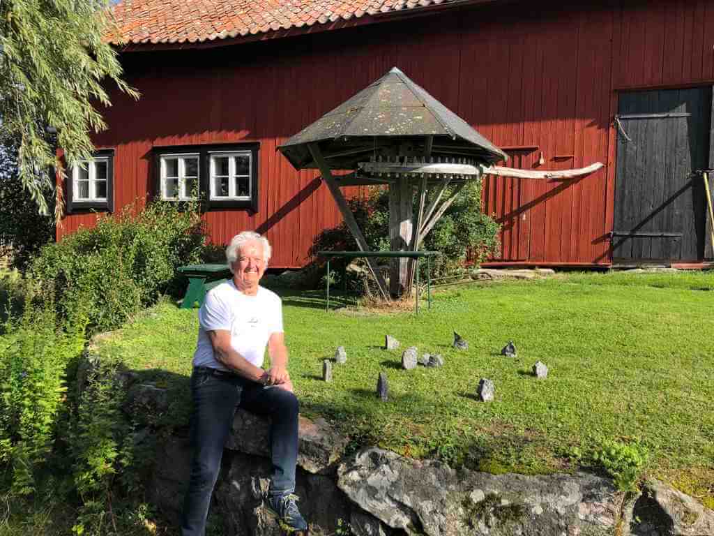 This is a colour photo of Neil, sitting on a low stone wall in front of a large, single-storey barn. The barn seems to be old - maybe 100 years old or more. It is of timber construction, with long, thin vertical boards painted in a terracotta colour. We can only see a section of the barn. On the far right is the closed left half of a solid, double barn-door entrance with long, horizontal metal hinges on the outside. The door seems to be unpainted and untreated, but weathered pine in a mid-grey colour. To the left of the dors is a large octagonal roof, about 3 m high. Underneath a curious inverted cone-shaped timber frame that may have been part of a mill but looks as if it no longer functions. Between the camera and the wall of the barn is a distance of about 15m covered in a green grass lawn. In the front half of the lawn closest to us and in front of the strange timber construction is a modern day replica of an ancient stone circle. It consists of eight stones on the circumference and three stones in the middle. The stones are roughly 150mm tall and 100mm x 100mm in section. They are all a grey granite and show no sign of having been worked. Neil is sitting on the wall to the left of the stone circle. Behind him on the grass is a green-painted wooden bench and above his head is a double casement, four-paned wooden window painted white in a black-painted frame. To its left is a matching single window and frame.