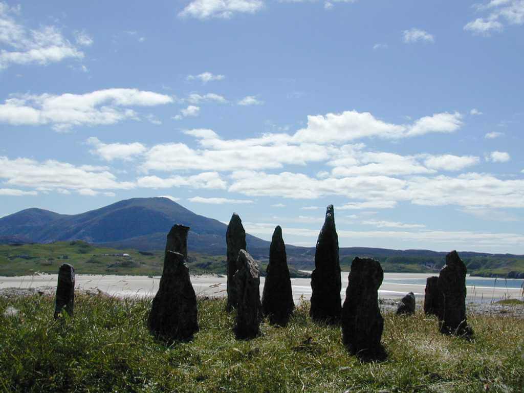 This is a colour photo of standing stones. The stones are a dark grey colour - probably granite and are standing on a grass bank. The other side of the bank is a wide, empty, large, pale, sandy beach which seemes to be the head of an inlet. The calm sea is visible to the right of the stones and behind the right-hand stones. Behind the beach is grass covered land in the distance and low mountains beyond. It is a bright, sunny day. The sun is infront of us but high and out of shot. The sky is a pale, watery blue and there are quite a few fair weather white clouds.