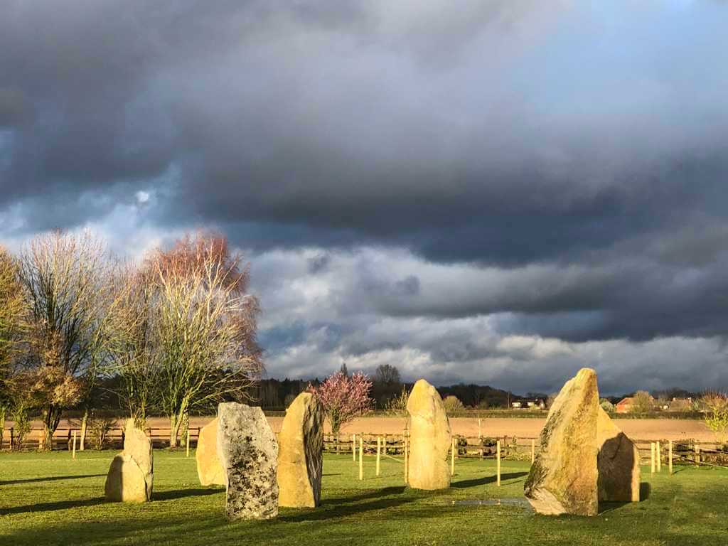 The Sussex Stone Circle. Group of upright 1.5m stones in a mown green grass field, 6 in a circle and one in the centre. Sun.