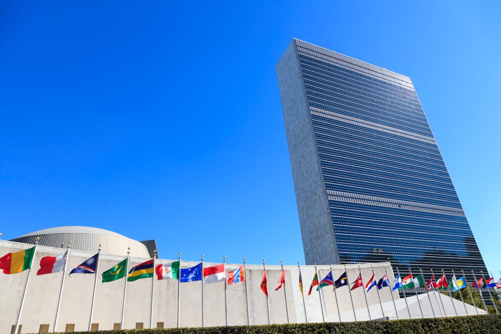 The United Nations Headquarters in New York, flags flying in a row in front. A brilliant blue sky behind and bright sunshine.